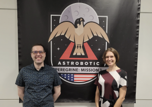 Image of two people standing in front of a large mission patch with a bird headed to the Moon